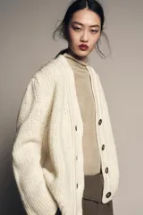 Chaqueta de punto de lana con cuello en pico, manga larga y cierre frontal con botones oscuros. Presenta un diseño de tejido grueso y holgado.