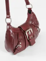 Cartera de hombro estilo baguette en color marrón con acabado brillante. Presenta un diseño con frunces, una hebilla central decorativa y dos hebillas laterales más pequeñas, todas con herrajes metálicos plateados.