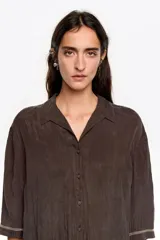 Camisa corta de corte boxy en color marrón, con cuello camisero, cierre frontal de botones y mangas 3/4. Presenta pespuntes decorativos a contraste en los puños y una textura sutil en el tejido.