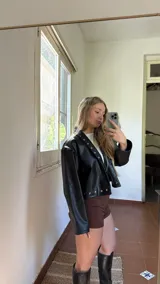 Campera corta de ecocuero negro, con diseño de solapas rectas decoradas con tachas metálicas plateadas a lo largo de los bordes frontales y el ruedo. Presenta mangas largas de corte amplio y estilo moderno.