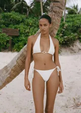 Top de bikini blanco con corte triangular, breteles finos que se atan al cuello y a la espalda, y aro dorado en el centro del busto.