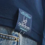 Remera de algodón azul marino con cuello redondo y mangas cortas. Presenta el logo de Adidas Trefoil bordado en blanco en el lado derecho del pecho y el escudo de la selección alemana de fútbol en el lado izquierdo. Incluye las icónicas tres tiras de la marca en color celeste claro a lo largo de los hombros y mangas.