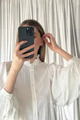 Camisa negra de corte clásico con cuello alto tipo mao con volados, cierre frontal de botones y mangas largas abullonadas con puños ajustados.
