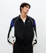 Campera deportiva de poliamida con diseño de bloques de color en negro, blanco y violeta. Presenta cuello alto, cierre frontal con cremallera, puños y ruedo elásticos, y forro interior de poliéster.