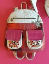 Mochila de cuero de diseño multicolor con detalles pintados a mano, solapa superior texturizada en blanco, bolsillos frontales con solapa fucsia y herrajes metálicos.