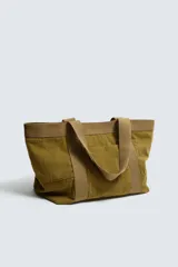 Bolso tipo shopper de gran capacidad, confeccionado en tejido con textura de pana. Cuenta con dos asas de mano resistentes, un bolsillo frontal plano y cierre principal mediante botón a presión. El interior incluye un bolsillo con cierre de cremallera.