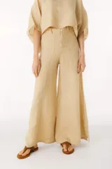 Pantalón ancho de tiro alto, color beige claro, confeccionado en lino. Presenta costuras verticales marcadas en el frente de cada pierna y cierre frontal con botón.