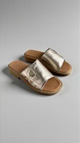 Sandalias planas de cuero color dorado pálido con detalle de arte pintado a mano en el lateral, logo metálico de la marca y base ergonómica.