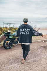 Hoodie de corte boxy fit en color negro con efecto lavado. Presenta estampado gráfico en la espalda con la frase 'Glassy Waves' en tipografía estilo fuego y detalles de llamas en las mangas.