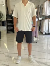 Camisa de manga corta color blanco marfil, confeccionada en tejido texturizado con cuello camisero, cierre frontal de botones y un pequeño bordado oscuro en el pecho.
