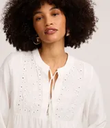 Blusa bata blanca de manga larga, confeccionada en viscosa y poliéster, con cuello padre, lazo y botones frontales, y detalles de bordado floral y broderie en el busto.