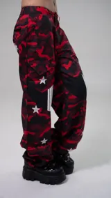 Pantalón de tiro bajo con estampado de camuflaje en tonos rojos y negros. Presenta diseño holgado, detalles de estrellas blancas aplicadas, paneles laterales con franjas verticales y bolsillos cargo.