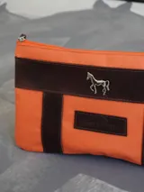 Sobre o cartuchera de lona fucsia con terminaciones y detalles de cuero marrón. Cuenta con cierre superior y aplique metálico de caballo en la parte frontal, además de una etiqueta de cuero con la marca grabada.