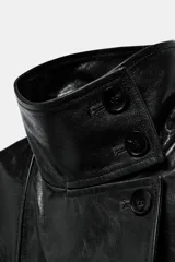 Trench de corte largo confeccionado en piel 100% con acabado brillante. Presenta cuello alto, hombreras marcadas, manga larga, bolsillos con solapa en el delantero y cierre frontal con botones. Incluye un cinturón ajustable del mismo material para entallar la silueta.
