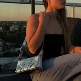 Cartera plateada tipo baguette con correa fina al hombro, detalles de tachas y cierres decorativos.