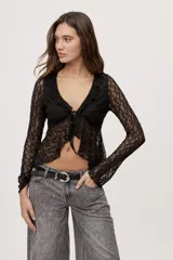 Top de encaje negro semitransparente con escote en V profundo, volados en los bordes y mangas largas. Cuenta con cierre frontal con lazos para anudar y diseño irregular.