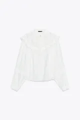 Blusa blanca de manga larga con cuello redondo, confeccionada con detalles de bordados y volantes a tono. Presenta cierre frontal con botones ocultos y puños ajustados.