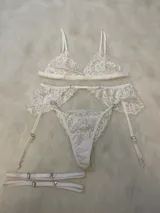 Conjunto de lencería de encaje blanco compuesto por un bralette triangular, una tanga y un portaligas con volados en la cadera y ligas integradas.