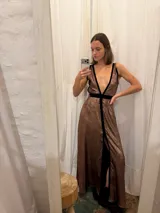 Vestido largo de seda color bronce con acabado brillante, escote profundo en V y espalda abierta. Presenta detalles en terciopelo negro que enmarcan el escote, recorren el frente y definen la cintura con una faja ajustable. Posee breteles anchos y una abertura frontal en la falda.