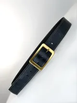 Cinto de cuero negro con textura granulada, costuras decorativas en los bordes y hebilla circular metálica en acabado plateado mate.