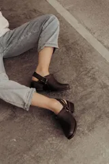 Zuecos de cuero color marrón oscuro, con diseño cerrado en la parte delantera, tira ajustable con hebilla metálica en el talón y taco cuadrado bajo.