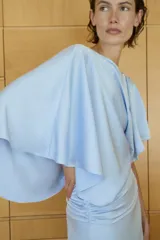 Vestido midi de tejido satinado color celeste, con cuello barco y diseño asimétrico con mangas tipo capa. Presenta un detalle drapeado en la cintura y abertura en la espalda.