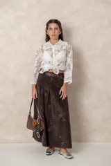 Camisa de organza con bordados florales, cuello camisero, cierre frontal con botones y mangas largas. Presenta un diseño traslúcido y ligero.