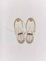 Championes de cuero color blanco con puntera y suela de goma marrón claro. Presentan detalles bordados dorados en los laterales y cordones anchos en color crudo.