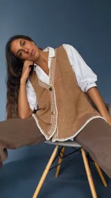 Chaleco tejido color tostado con destellos de lurex, escote en V y cierre frontal con botones. Presenta detalles de bordados y terminaciones en crochet en color crudo.