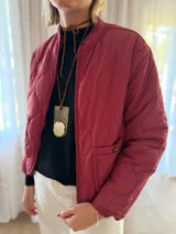 Campera tipo bomber acolchada con diseño de pespuntes ondulados, cierre frontal y bolsillos laterales con botón. Presenta un corte moderno y acabado en color bordo.