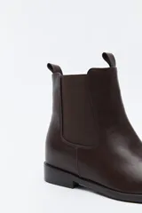 Bota tipo chelsea de caña media confeccionada en cuero sintético, con paneles laterales elásticos y presillas superiores. Presenta un diseño minimalista con suela plana y punta redondeada.