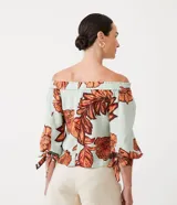 Blusa celeste con estampado de hojas color naranja y marrón, con escote hombro a hombro, mangas 3/4 con lazos en los puños y corte holgado.