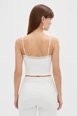 Musculosa de punto blanco, corte crop, con tirantes finos, escote en V adornado con detalles de encaje y un pequeño lazo en el centro.