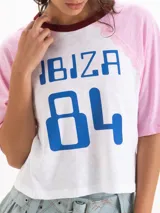 Remera estilo raglán de manga corta, con cuerpo blanco, mangas rosadas y cuello redondo bordó. Cuenta con un estampado frontal en azul con las palabras 'IBIZA 84' y detalles de encaje en las costuras de las sisas.