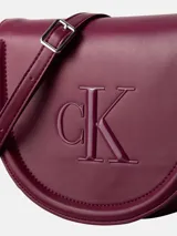 Cartera saddle Calvin Klein de diseño estructurado en color bordó, con solapa superior, cierre magnético, tirante ajustable para el hombro y logo monograma grabado en el frente.