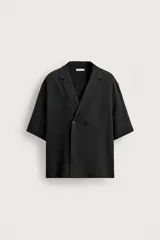 Camisa de manga corta color negro, de corte relajado, con cuello con solapas de muesca y cierre frontal de botonadura cruzada.