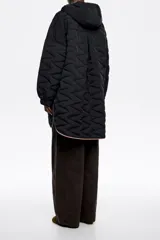 Abrigo largo negro de corte línea A, confeccionado en tejido técnico acolchado con patrón de costuras en zigzag. Posee cuello subido con capucha integrada y cordones a contraste, cierre frontal de botones de presión ocultos, bolsillos de vivo y puños anchos elastizados.