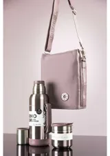 Bolso tipo bandolera color marrón, de diseño rectangular y minimalista. Cuenta con una correa larga ajustable con el nombre de la marca grabado y un aplique circular con el logo en la parte inferior frontal.