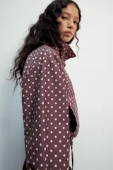 Cazadora negra con estampado de lunares blancos, cuello subido, manga larga, bolsillos delanteros, bajo ajustable con cordones y cierre frontal con cremallera y solapa con botones ocultos.