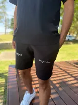 Bermuda deportiva negra de felpa, con bolsillos y cordón ajustable en la cintura. Presenta el texto "TOP STYLE SINCE 2021" estampado en blanco en la pierna derecha y "follow your dreams" en cursiva en la pierna izquierda.