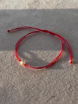Pulsera de hilo rojo regulable con cierre de nudo corredizo y un punto de luz central engarzado en oro laminado.
