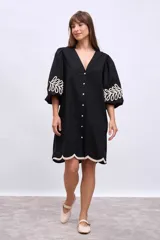 Vestido corto negro con escote en V, mangas 3/4 abullonadas y cierre frontal con botones. Presenta apliques bordados en color crudo en mangas y ruedo.