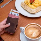 Billetera tarjetero automática con estructura de aluminio y revestimiento de cuero bicolor en tonos verde oliva y beige. Cuenta con un cierre de broche a presión y el logo de la marca grabado en la parte inferior.