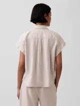 Camisa de lino blanca de manga corta, con cuello camisero, cierre frontal de botones y corte corto.