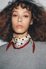 Mini bandana de forma triangular confeccionada en viscosa satinada. Presenta un diseño estampado con lunares negros sobre fondo crema, con bordes en color rojo y detalles de líneas geométricas negras.