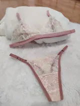 Conjunto de lencería compuesto por bralette y tanga regulable, confeccionado en encaje color champagne con detalles florales bordados en hilo rosado y terminaciones en cinta color rosa viejo.