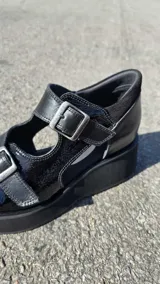 Sandalias de cuero color negro, de estilo cerrado con aberturas laterales, plataforma y doble tira ajustable con hebillas metálicas.