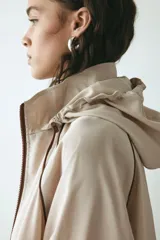 Campera rompevientos de color beige, modelo oversized con cuello alto y capucha con cordón ajustable. Cuenta con cierre frontal marrón, mangas raglán con puños elásticos, bolsillos laterales y detalles de ribetes en tono marrón oscuro.