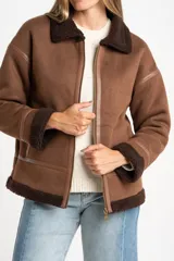 Chaqueta tipo gamulán de calce oversize en color marrón, con cuello y ribetes de corderito en contraste. Presenta cierre frontal de cremallera, bolsillos laterales y detalles de tiras decorativas en material sintético.