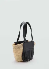 Bolso de mano tipo cesta confeccionado en tejido de fibra natural, con detalles en cuero negro que incluyen doble asa corta, solapa frontal y flecos decorativos.